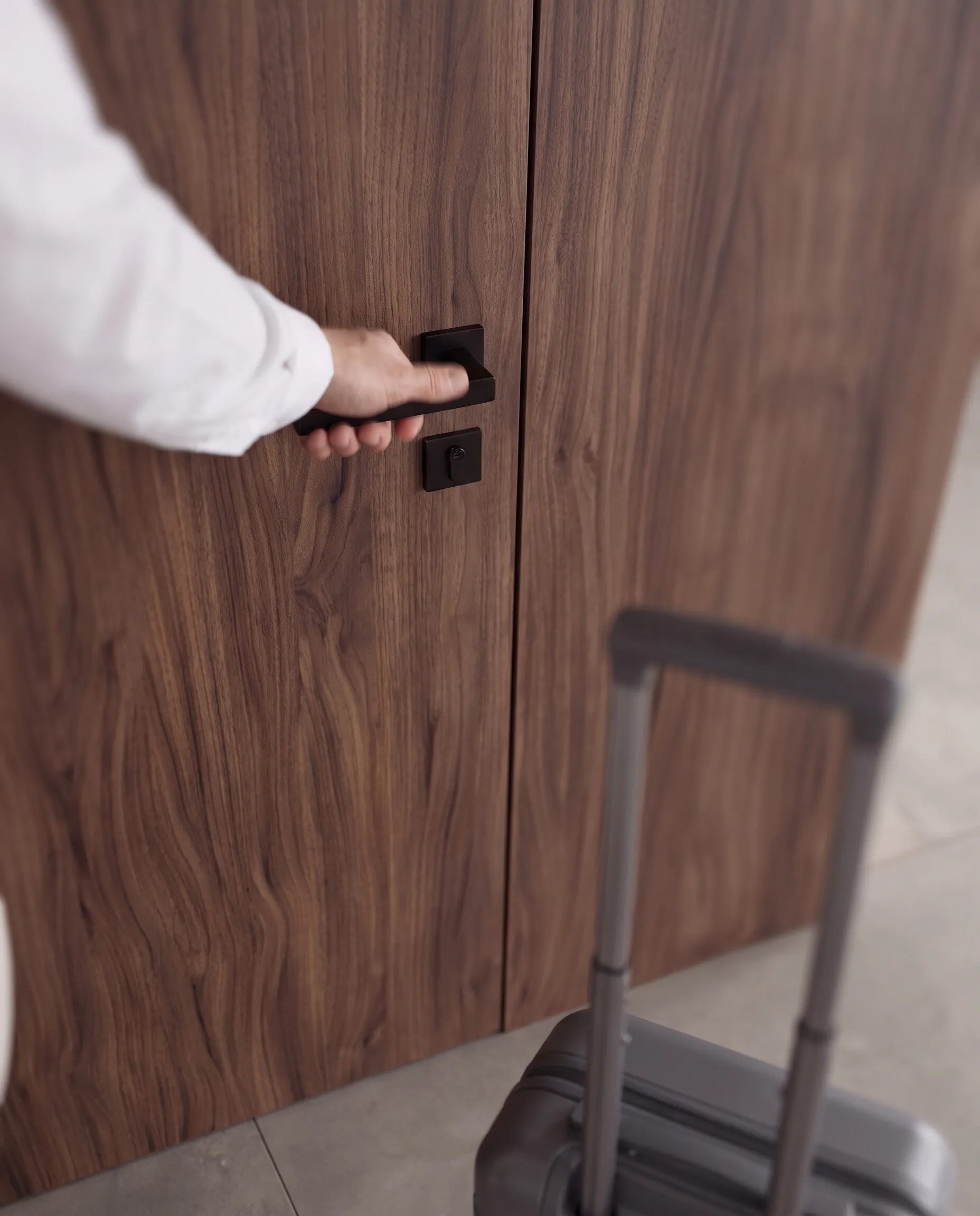 Entrance: a hand on the black handle of a walnut door, a suitcase beside it
